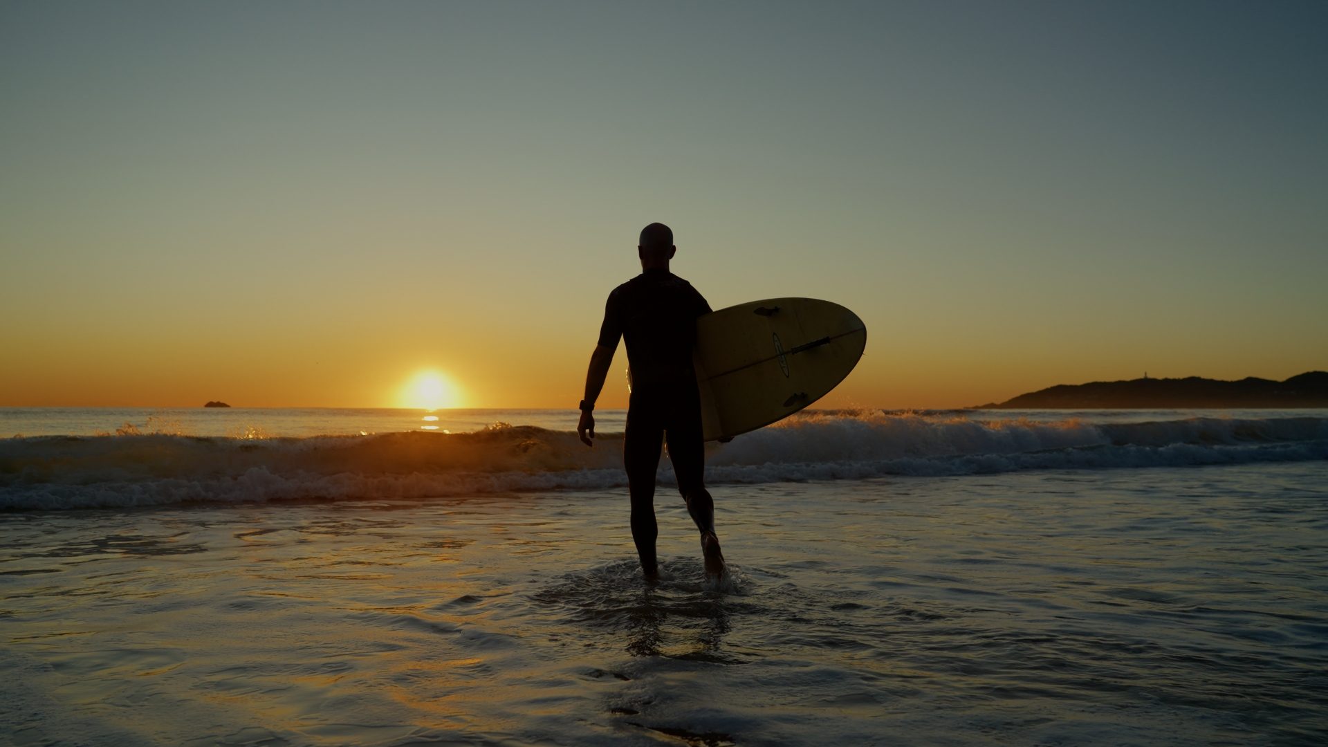 A surfer carrying a board out of the shallows at sunrise
