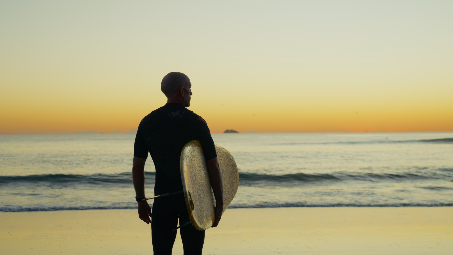 A surfer standing on the shore at dusk, holding a board